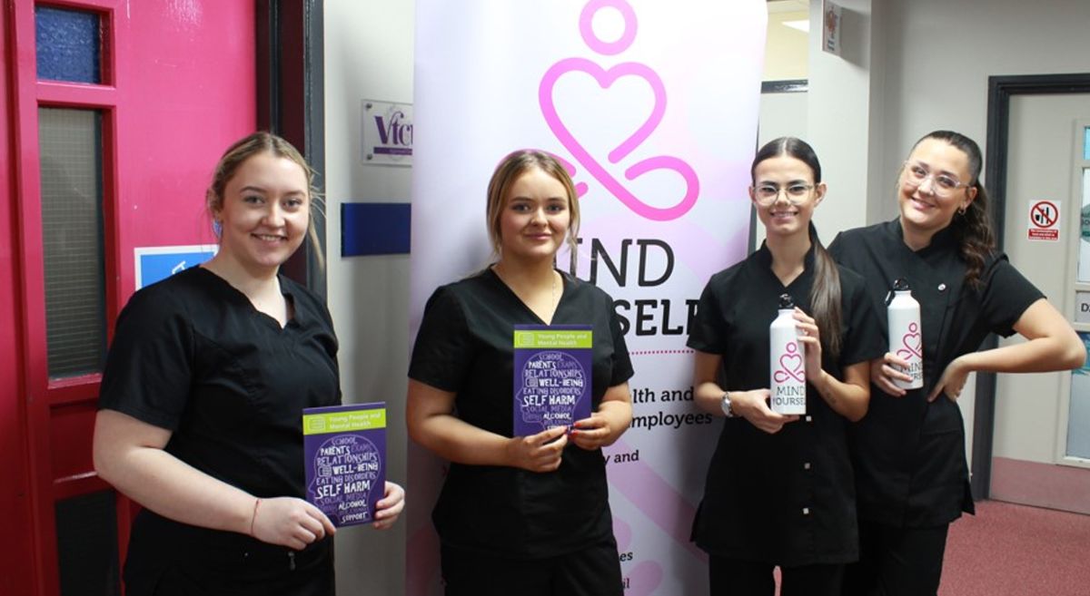 Four female Beauty Therapy students wearing dark salon uniform, holding wellbeing leaflets and water bottles with Mind Yourself pop up stand in backfground.